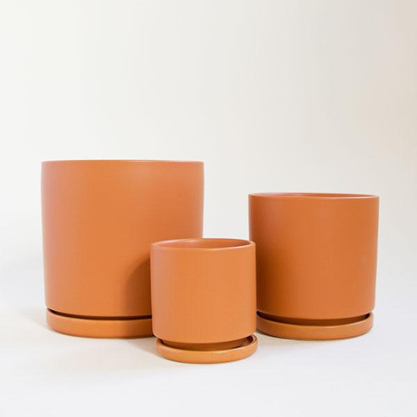 Three terracotta orange cylinder planters, each complete with matching water tray. The three sit empty staggered in front of each other in sizes small, medium and large.