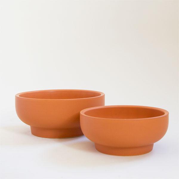 Two terracotta orange pedestal bowl planting pots, both vessels are round and have tapered bases. The one in front is the smaller of the two and the pair sit empty besides each other.
