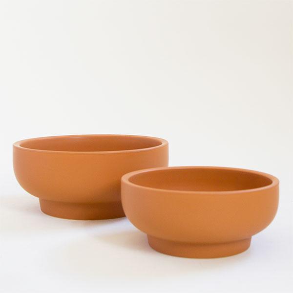 Two burnt orange pedestal bowl planting pots, both vessels are round and have tapered bases. The one in front is the smaller of the two and the pair sit empty besides each other.