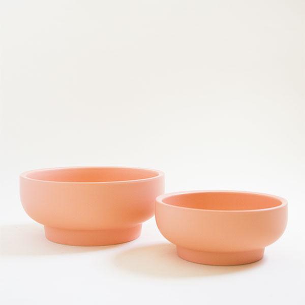 A pair of two salmon colored pedestal bowls, both with tapered bases. To the left is the larger of the pair and to the right sits a slightly shorter bowl. They both are positioned empty besides each other.
