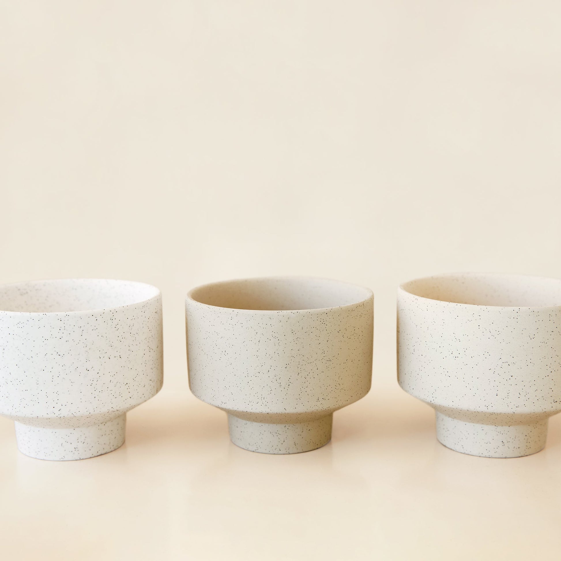 Three round, ceramic white pots with a tapered bottom. The pots have black speckles and are in an array of warm cream tones.