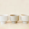 Three round, ceramic white pots with a tapered bottom. The pots have black speckles and are in an array of warm cream tones.