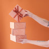 A stack of four salmon pink gift boxes with a magnetic closure and darker pink bird of paradise line graphic along with a rust colored ribbon tied in a bow, not included with purchase.