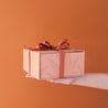 A salmon pink gift box with a magnetic closure and darker pink bird of paradise line graphic along with a rust colored ribbon tied in a bow, not included with purchase.