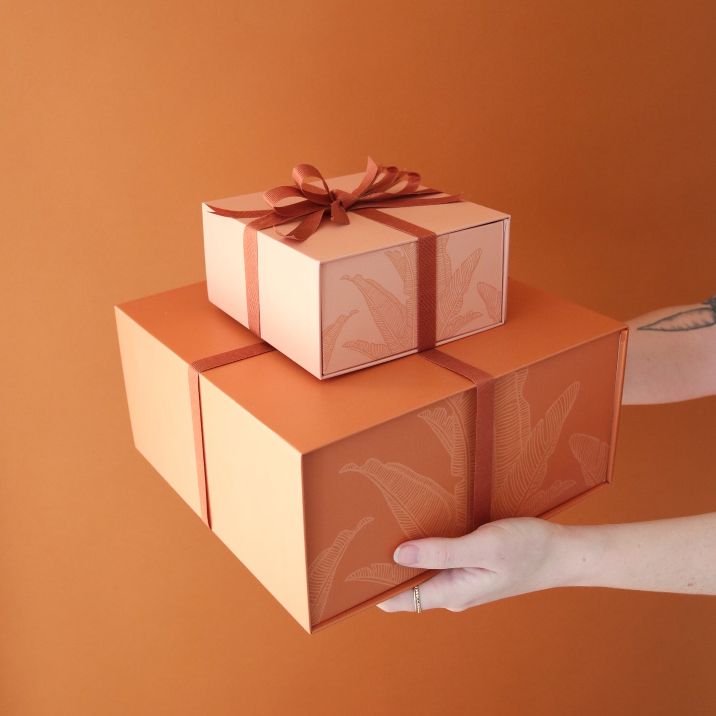 On an orange background is two different sized light orange gift boxes stacked on top of one another with white bird of paradise outline designs and wrapped with a rust colored ribbon.
