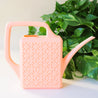 On a cream background staged next to a green pothos is a pink plastic watering can with a breeze block design on both sides along with a long spout and a squared off handle for easy watering.