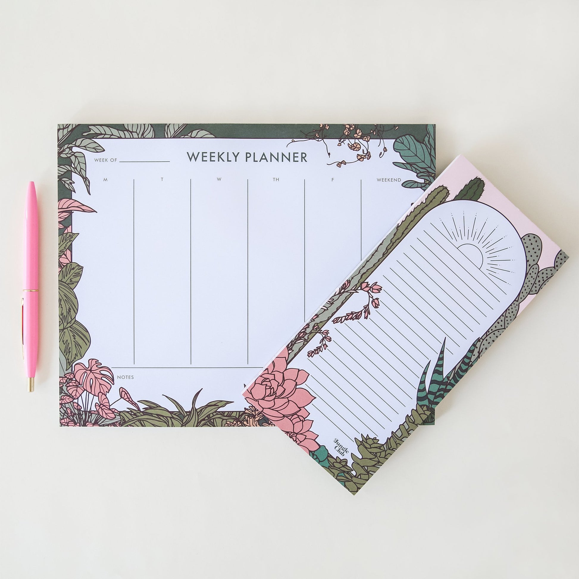 This weekly planner pad has a section for each day of the week with a deep green border, accented by an array of natural, soft toned jungle plants. Besides lays a rectangular notepad bordered with muted cacti and succulent plants. A baby pink ball point pen lays vertically to the left of the weekly planner. 
