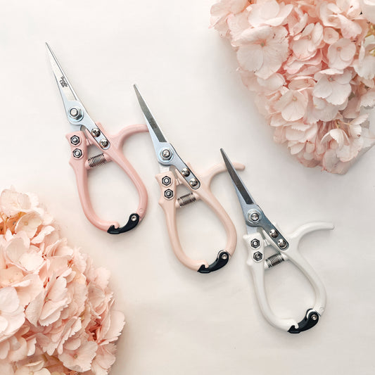 Pair of pruning shears with pastel pink handles and metal blades. The outer blade reads 'jungle club' in small, playful lettering. The pruning shears have a black clasp at the top of the handles to secure them closed. In this photograph, they're staged on a white background next to two different color ways of the same shears and in between light pink hydrangeas.