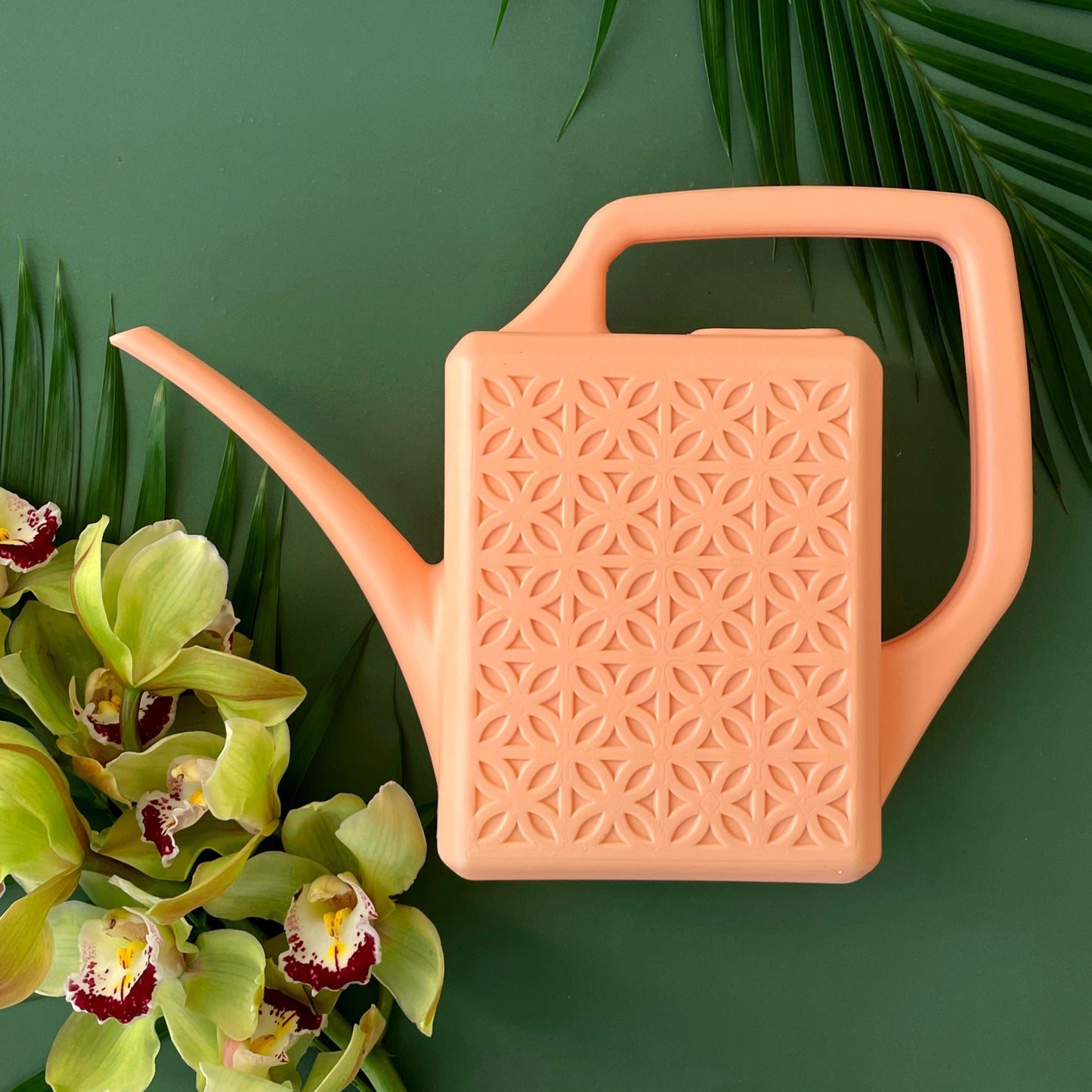 On a dark green background staged with tropical flowers and palm leaves is a light pink plastic watering can with a breeze block design on both sides along with a long spout and a squared-off handle for easy watering.