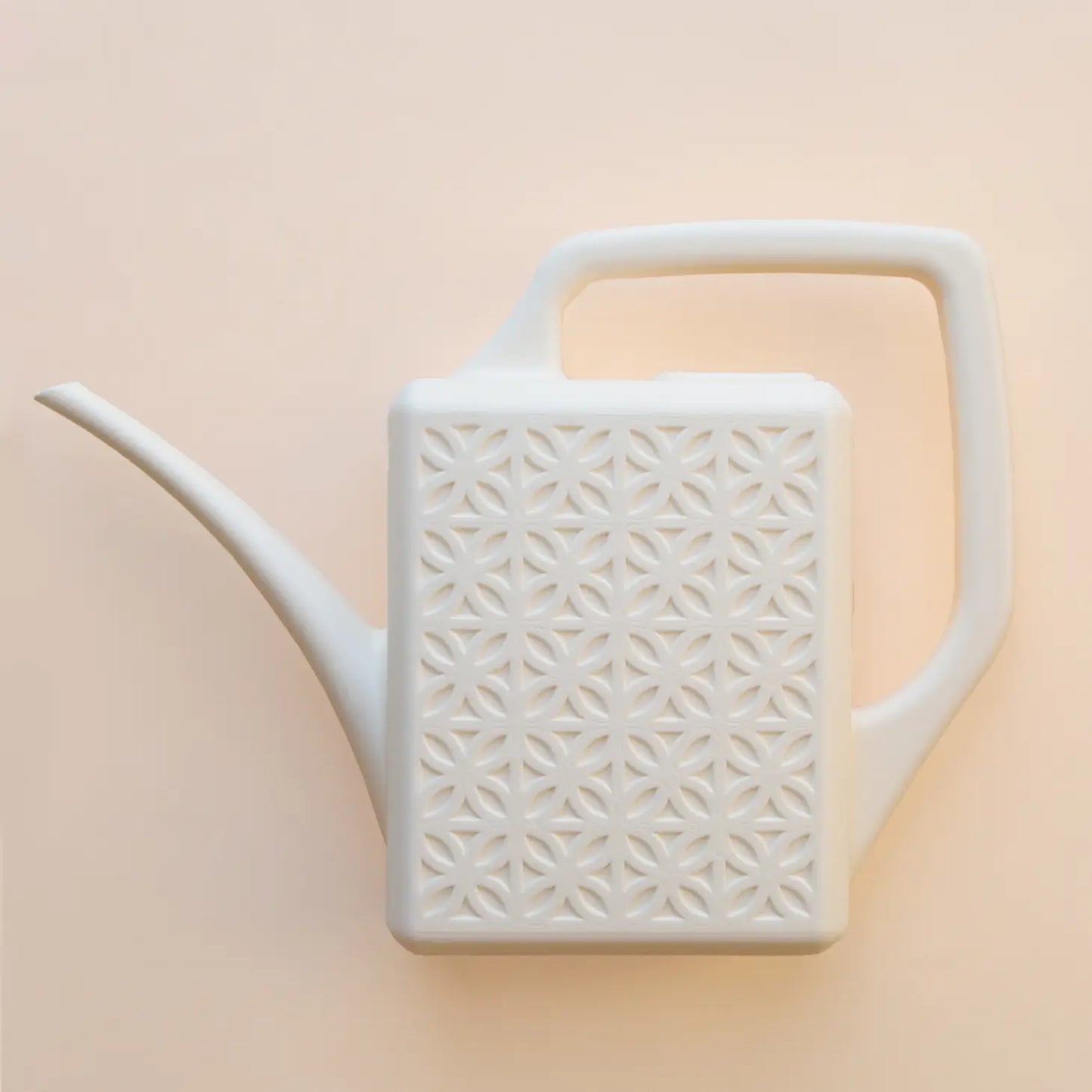 On a peachy background is a white plastic watering can with a breeze block design on both sides along with a long spout and a squared off handle for easy watering.